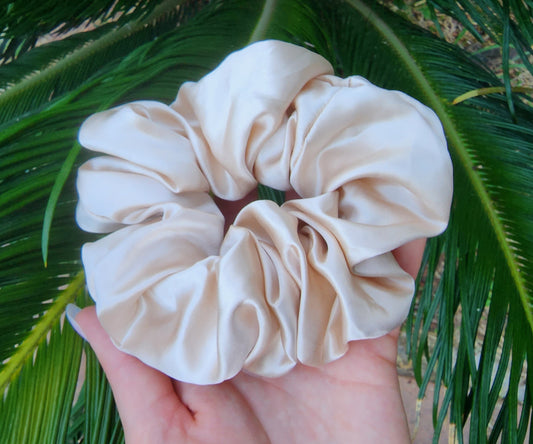 Oat Milk oversized mulberry silk scrunchie in beige cream held in a hand against green palm leaves