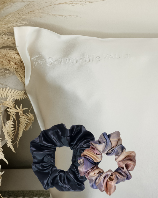 Silk Indulgence Pack – lifestyle image showing corner of 22 momme Mulberry Silk pillowcase with logo visible, classic printed scrunchie and oversized plain scrunchie in front.